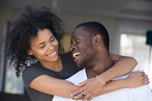 Portrait of happy black husband and wife embrace looking at each other, smiling African American couple cuddle having fun together, excited girlfriend hug boyfriend from behind, showing love and care